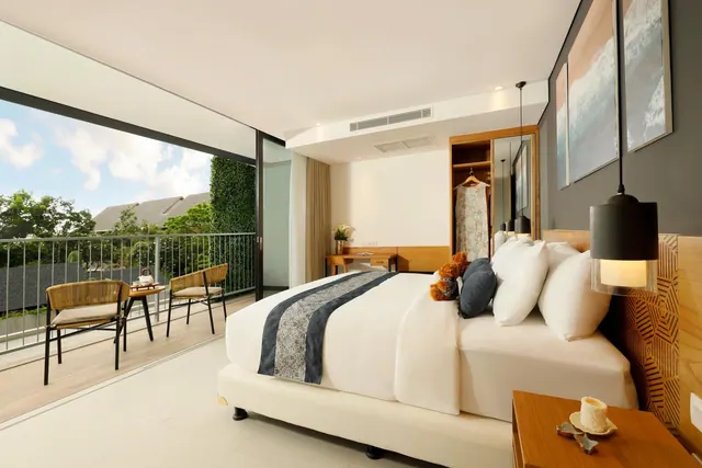 A bedroom with a made king bed, a wardrobe, a small work desk and a glass-door balcony with rattan chairs and a table, looking out onto trees.