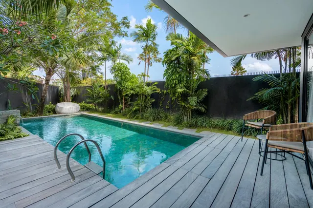 A private swimming pool with a grey timber deck, rattan chairs and tropical planting beside a dark garden wall.