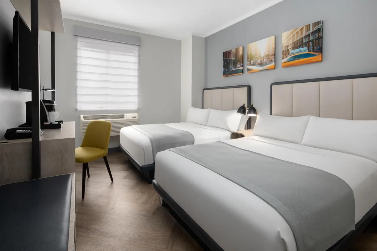 A bedroom with two double beds, a yellow chair, a wall-mounted television, a desk with a coffee machine, and framed city street prints on the wall.