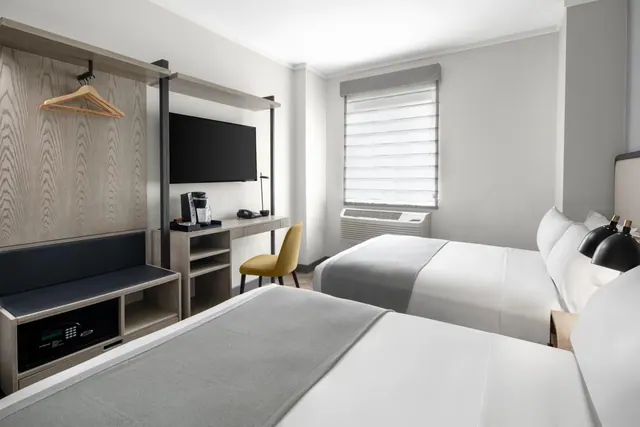 A hotel room with two double beds, a wall-mounted television, a desk with a coffee machine, a yellow chair, and a window with a wall air conditioner beneath it.