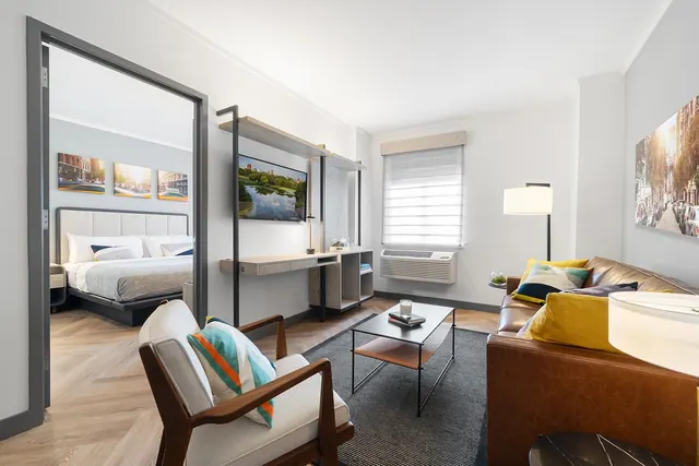 A living area with a leather sofa, armchair, coffee table, wall-mounted desk and television, with a bedroom and king bed visible through the doorway on the left.