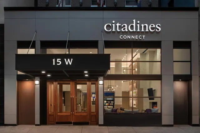 Night view of the Citadines Connect hotel facade at 15 W, with illuminated signage, wooden entrance doors and a glass-fronted lounge with sofas visible inside.