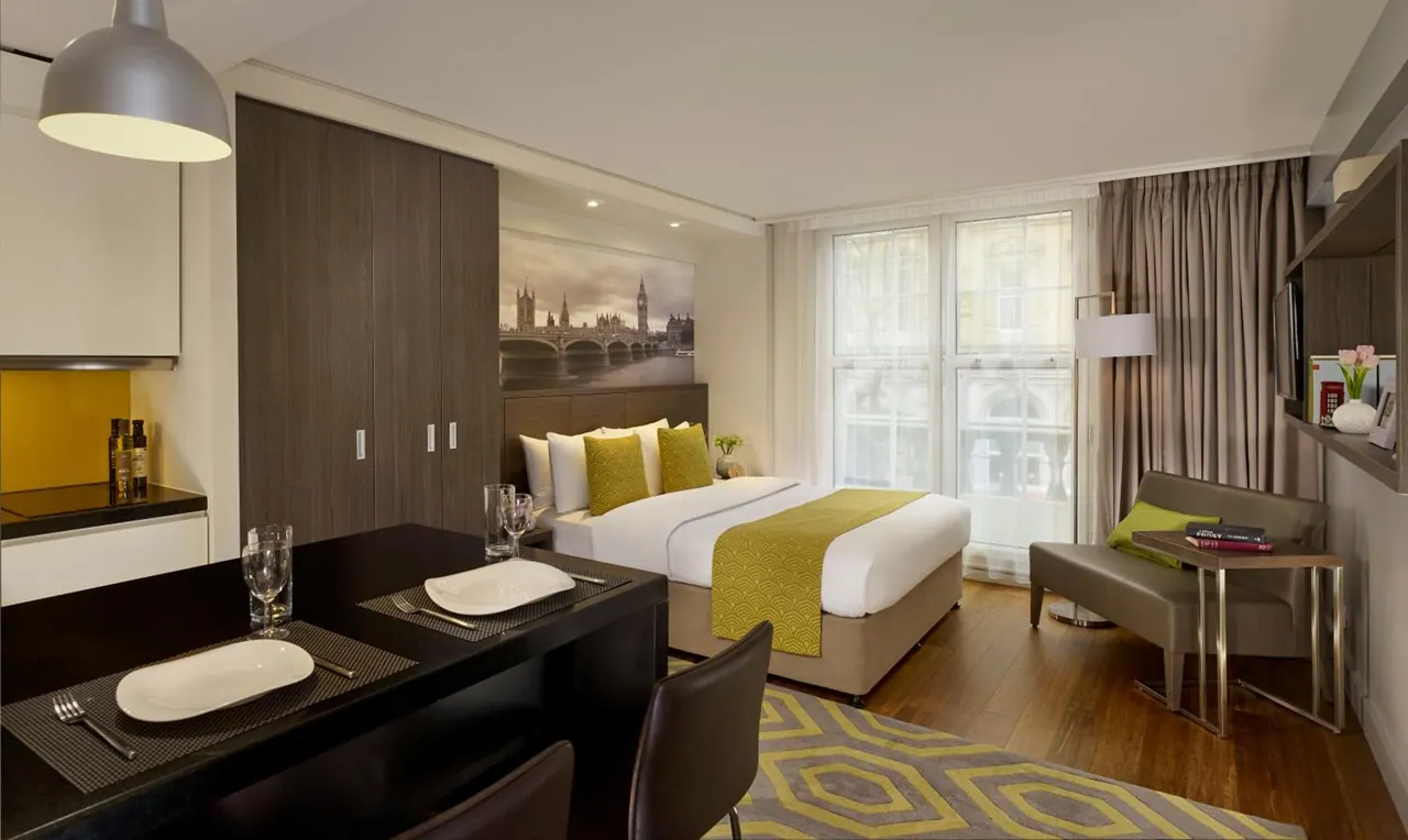 A studio room with a double bed, a dining table, a kitchenette and a wall mural of Big Ben and the Houses of Parliament above the headboard.