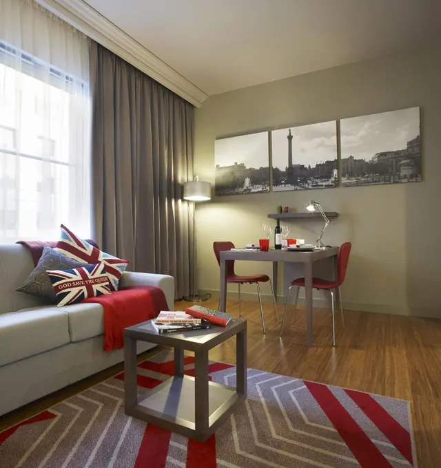 A living area with a grey sofa, Union Jack cushions, a red throw, a small table with two red chairs, and a black-and-white Trafalgar Square print on the wall.