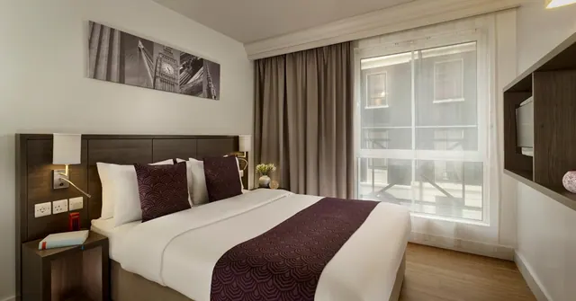 A double bed with a dark wood headboard and burgundy runner sits in a hotel bedroom, with a window looking onto a building across the way and a wall-mounted television at right.