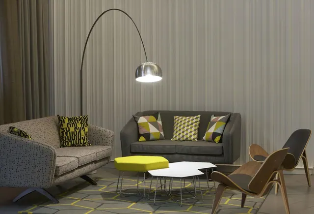 A lounge seating area with a patterned grey sofa, a dark grey loveseat, an arc floor lamp, a yellow ottoman, white hexagonal tables and wooden shell chairs on a geometric rug.