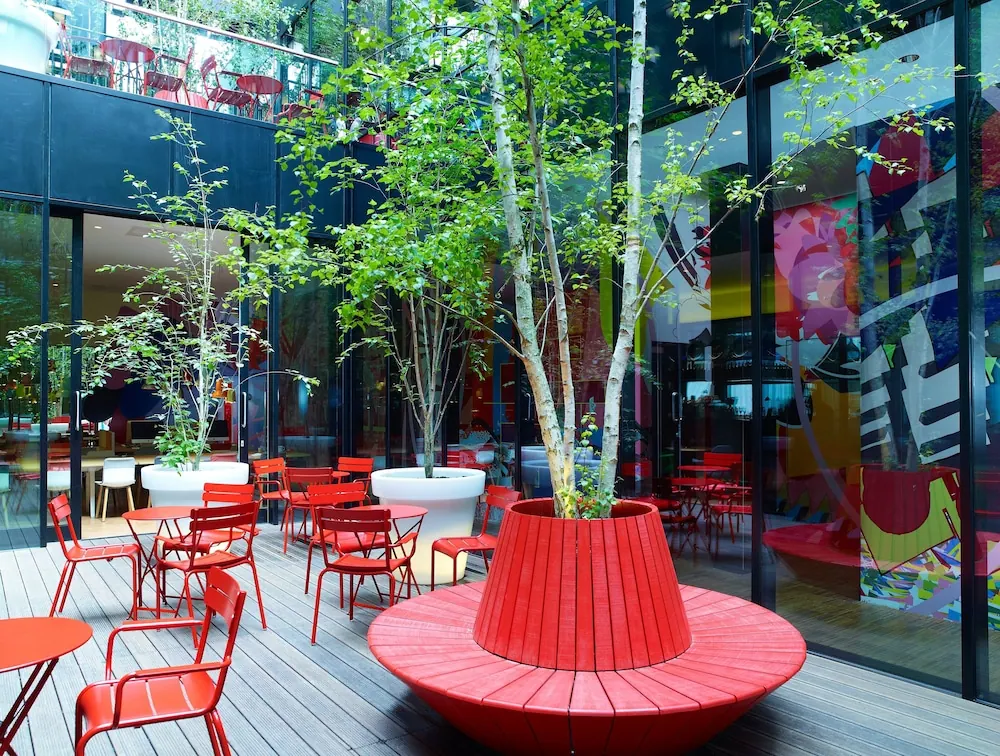 An open-air courtyard terrace with red metal tables and chairs, a red circular bench around a birch tree in a planter, and glass walls with colourful artwork.