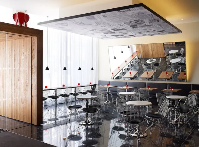 A lounge area with round tables, wire chairs, a dark banquette along the wall, a wall mirror and a newspaper-print ceiling panel.