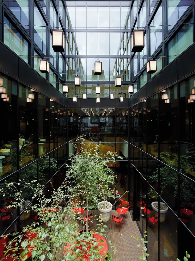 A glass-walled atrium with hanging square lanterns, potted trees, and red tables and chairs on a timber floor.