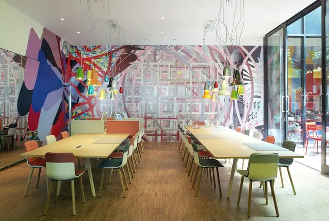 A dining room with long wooden tables, multicoloured chairs and pendant lamps, beside a large painted mural wall and floor-to-ceiling glass doors onto an outdoor area.