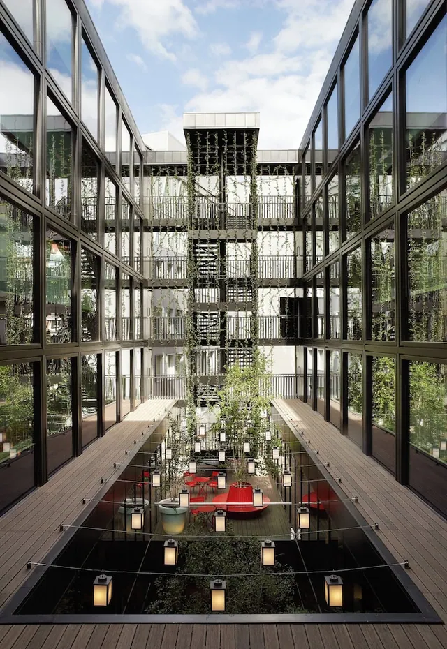 A glass-walled central courtyard with a long reflecting pool, lanterns, potted trees, red seating and an ivy-covered staircase tower.