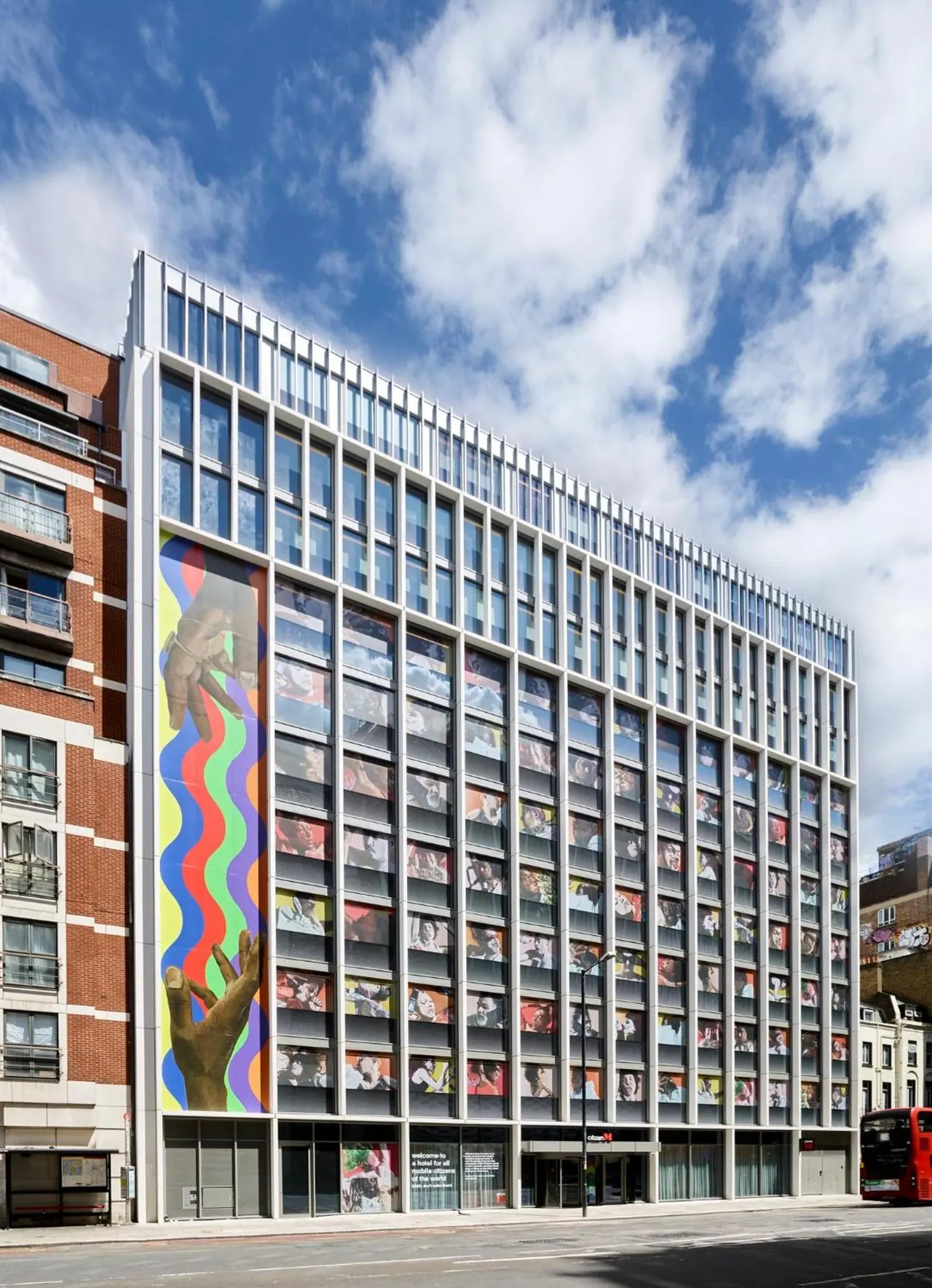 Exterior of a tall modern hotel building with a colourful wavy mural on its left side, a street in front, and a red bus at the right edge.