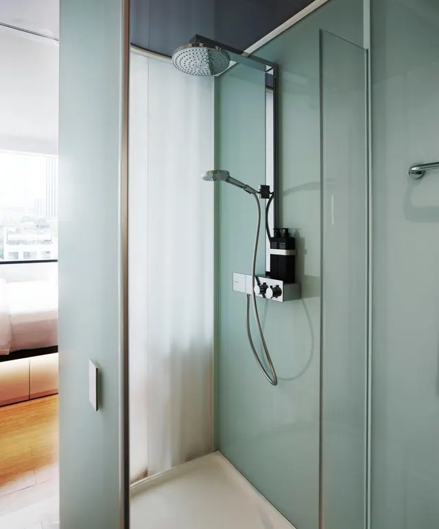 A glass-walled walk-in shower with a rain head, a handheld shower and wall-mounted controls, with a bedroom visible through the open doorway.