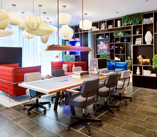 A lounge area with a long grey table, grey office chairs, red and blue sofas, pendant lamps, and black shelving holding decorative objects and plants.