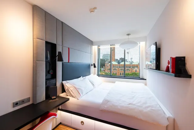 A small bedroom with a double bed, a wall-mounted television, a desk with a red chair, and a window looking onto a red brick building.