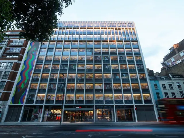 The exterior of a tall glass-fronted hotel building at dusk, with illuminated rooms, a street-level entrance and a red bus passing on the road.