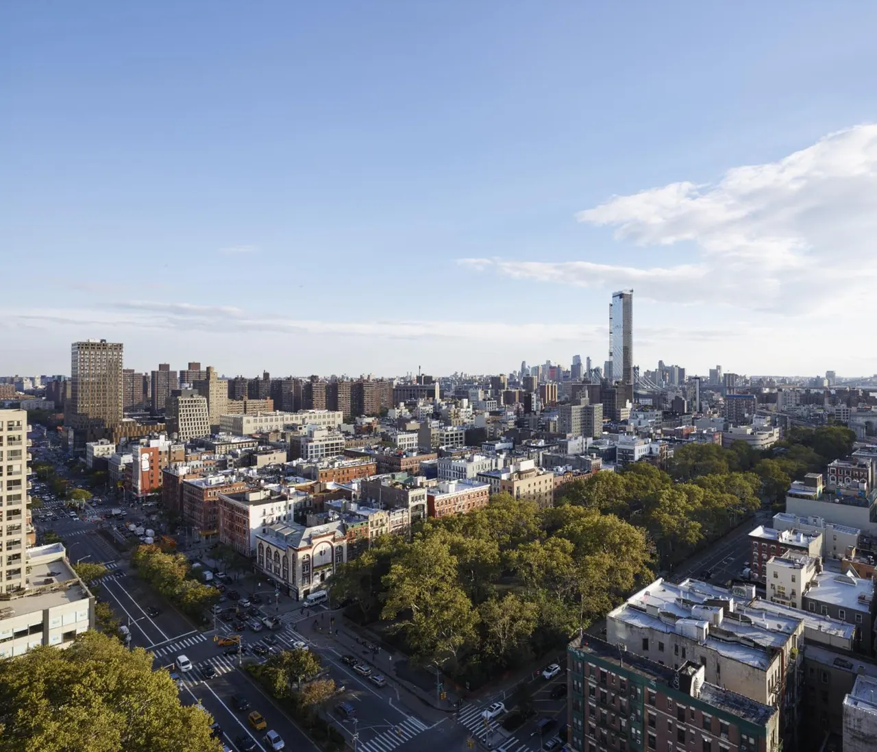 Elevated view over a dense city neighbourhood with a tree-filled park, low-rise brick buildings, and a high-rise skyline in the distance under a partly cloudy blue sky.