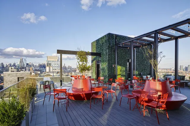 Rooftop terrace with red chairs and tables, a green wall, and a view over the city skyline including the Empire State Building.