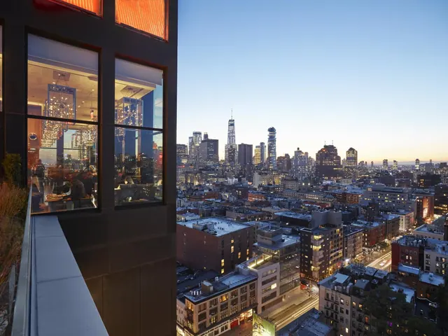 Dusk view over a dense city skyline including One World Trade Center, seen from a terrace beside a glass-fronted restaurant with chandeliers.