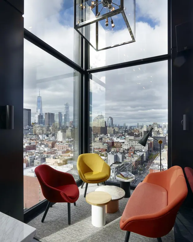 A corner lounge with floor-to-ceiling windows holds four colourful armchairs and side tables, overlooking a city skyline with One World Trade Center visible.