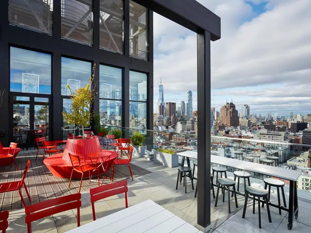 A rooftop terrace with red chairs, high bar tables and stools, and a view over city buildings including One World Trade Center.