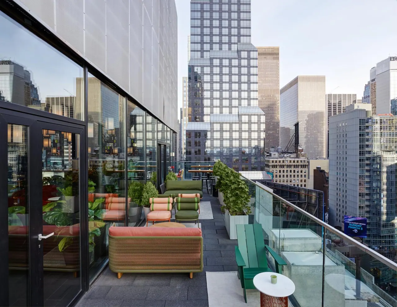 A rooftop terrace with outdoor sofas, armchairs and planters beside a glass railing, with tall city office buildings in the background.