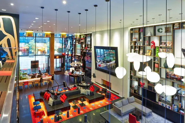 A double-height lobby lounge with a sunken seating area on a red star-patterned carpet, bookshelves, pendant lamps and a wall-mounted television, with a street view through the glass front.