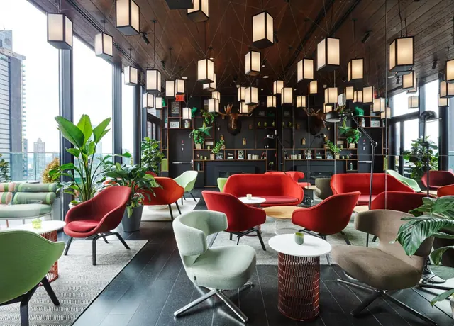A lounge with red and green armchairs, low round tables, hanging lantern lights and a mounted moose head on the wall, with tall city buildings visible through the windows.