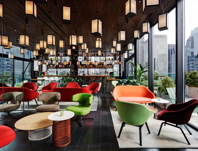 A lounge with red, orange and green armchairs and sofas beneath rows of square pendant lights, with a bar at the back and floor-to-ceiling windows showing city buildings.