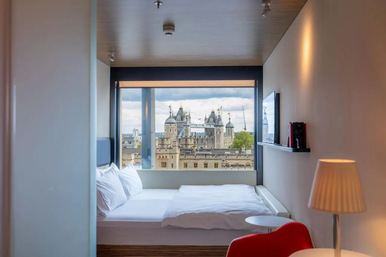 A small bedroom with a double bed beneath a large window showing Tower Bridge and the Tower of London, with a red chair and a lamp in the foreground.