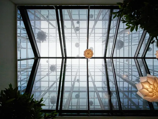 Looking up at a glass-paned ceiling with a dark metal frame, hanging paper lanterns and green plants at the edges.