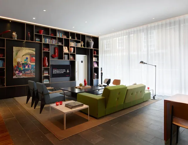 A lounge with a green modular sofa, grey armchairs, a coffee table, a wall of black shelving with a television, and tall windows behind sheer curtains.