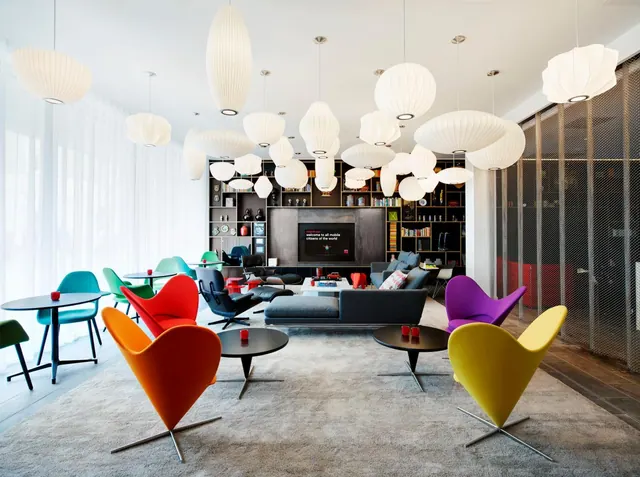 A lounge area with brightly coloured heart-shaped armchairs, white pendant lamps, grey sofas, a low table, and a wall-mounted television in front of bookshelves.