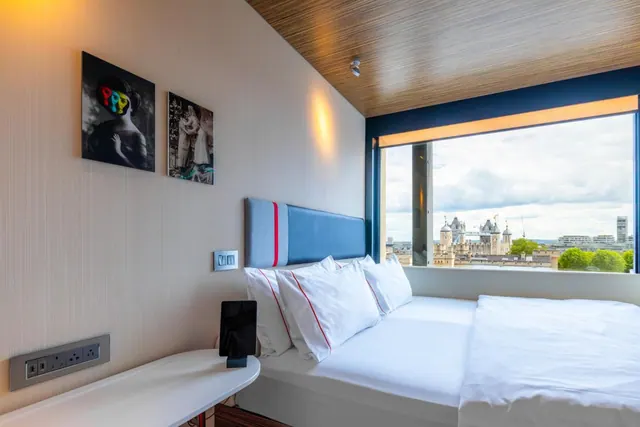 A double bed with white linen and a blue headboard sits beside a large window with a view of the Tower of London.