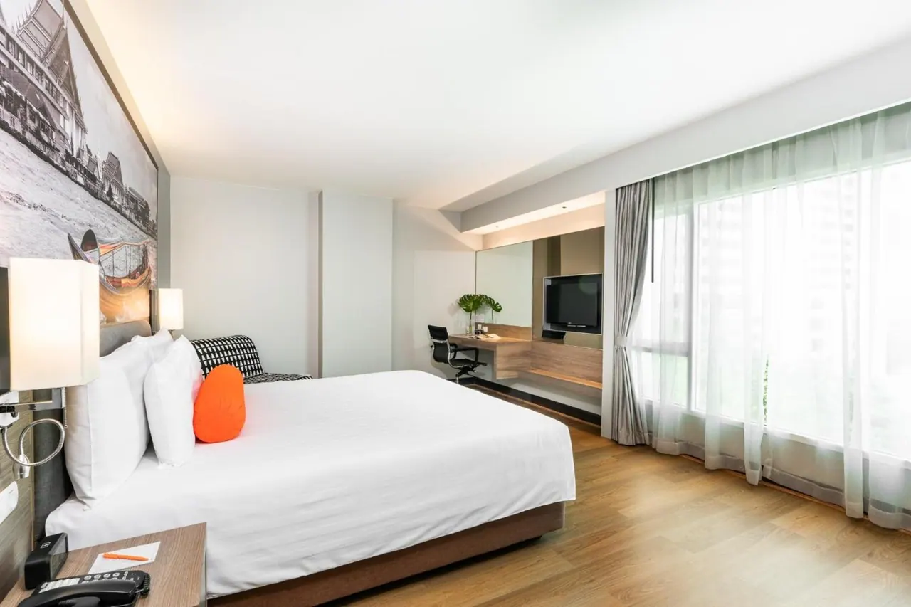 A bedroom with a made king bed, a framed black and white photo above the headboard, a work desk with a wall-mounted television, and floor-to-ceiling windows with sheer curtains looking out over city buildings.