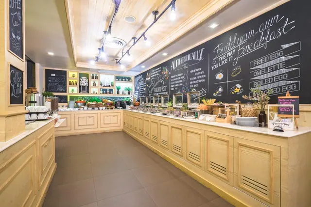 A breakfast buffet counter with warming trays runs along a wooden cabinet beneath a chalkboard wall that reads Build your own breakfast, under ceiling track lights.