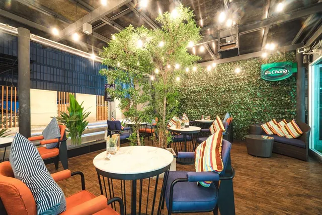 A lounge area with orange and navy armchairs, round marble-top tables, a tree under string lights, a green leaf wall and a rattan sofa.