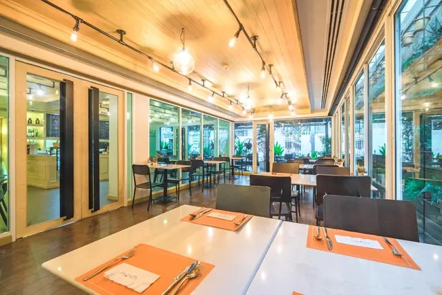 A restaurant dining room with set tables, dark chairs, a wooden ceiling with track lights, and floor-to-ceiling windows looking out onto plants and a building.