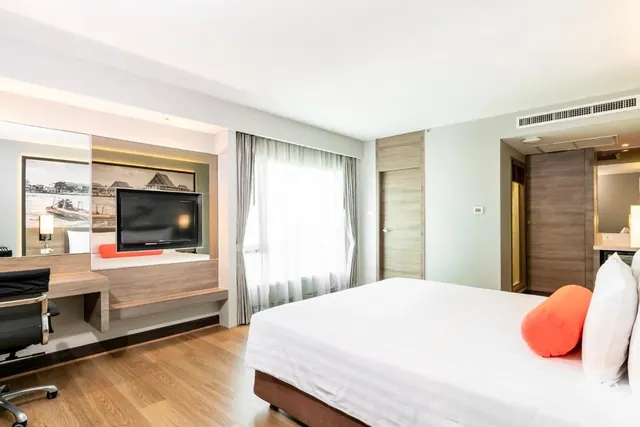 A bedroom with a large white bed with orange cushions, a wall-mounted television above a built-in unit, a desk with an office chair, and a wooden wardrobe beside the door.