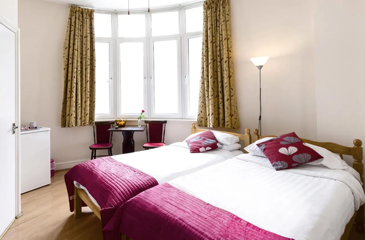 A twin-bedded hotel room with a bay window, a floor lamp, a small table with two chairs, and a small white fridge by the door.