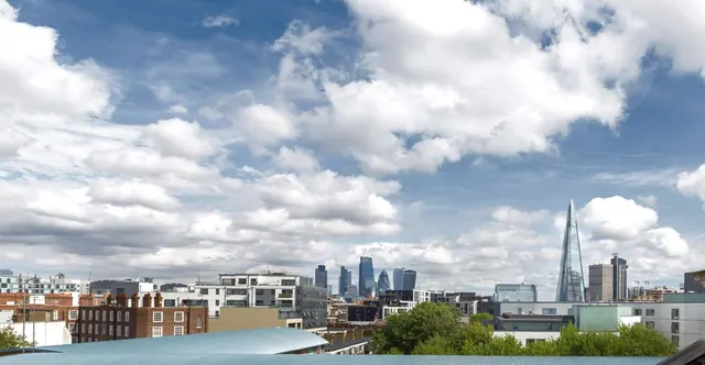 A view over London rooftops and office towers toward The Shard under a cloudy blue sky, with a curved metal roof in the foreground.