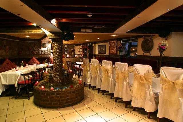 A restaurant dining room with a pebble-clad pillar and water feature in the centre, laid tables on the left, and rows of white chair covers with gold sashes on the right.