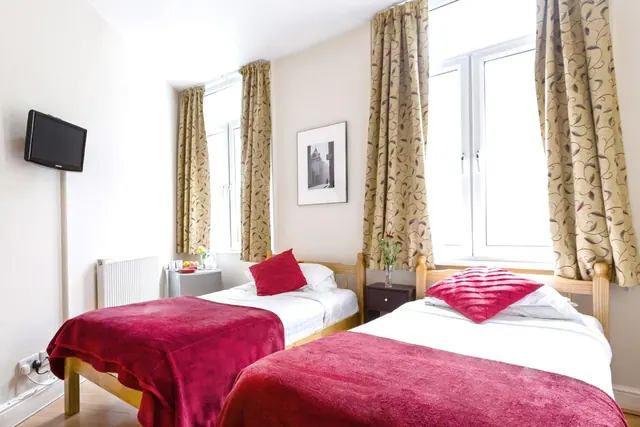 A bedroom with two single beds with red throws, a wall-mounted television, patterned curtains and a radiator.