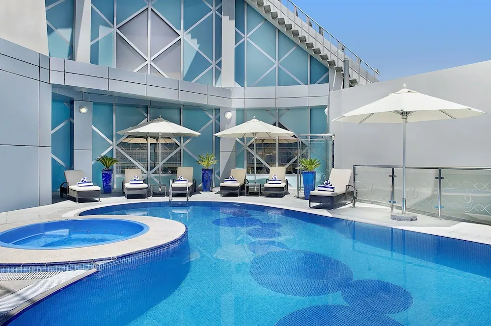 A rooftop swimming pool with a round hot tub, white parasol umbrellas and sun loungers with folded blue towels, set against a blue glass building.