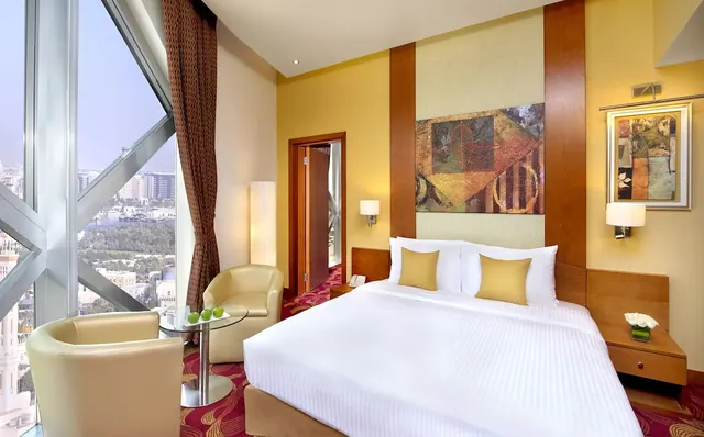 A hotel bedroom with a large bed, two armchairs beside a round table, and angled floor-to-ceiling windows looking over a city.