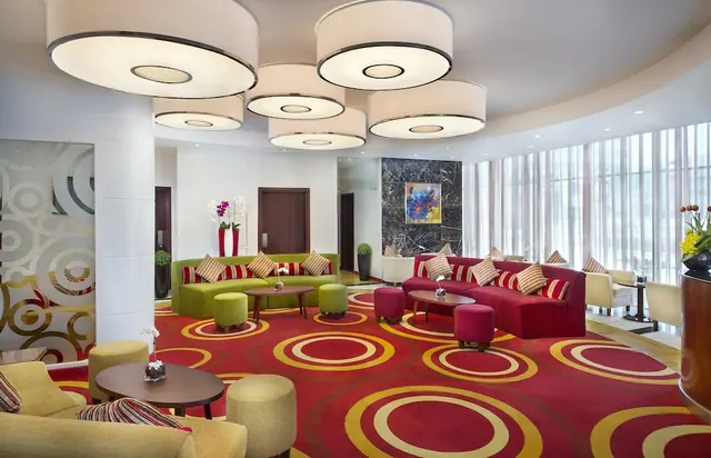 A lounge seating area in a hotel lobby with green and red sofas, round wooden tables, and a red patterned carpet under large drum pendant lights.