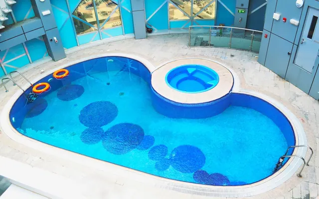 An indoor kidney-shaped swimming pool with a round hot tub attached, two orange life rings on the left edge, and blue glass walls behind.