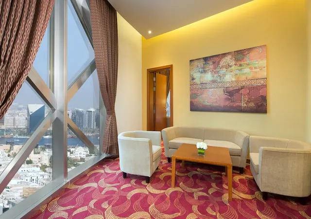 A seating area with two armchairs, a sofa and a low wooden table sits beside floor-to-ceiling windows overlooking a city and a waterway.