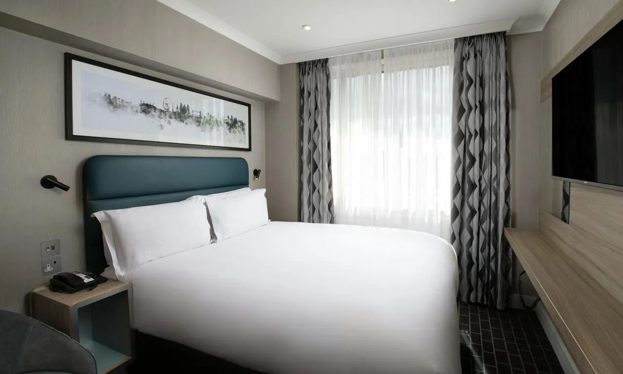 A made-up double bed with a teal headboard, a bedside table with a telephone, a wall-mounted television and patterned curtains beside a window in a hotel bedroom.