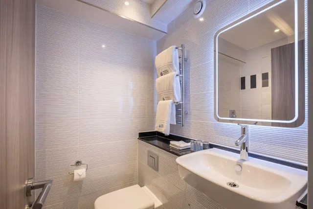 A bathroom with a backlit mirror above a white wall-hung sink, a toilet, a heated towel rail holding white towels, and textured wall tiles.